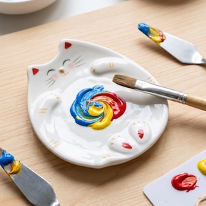 May include: A white ceramic cat-shaped paint palette featuring a painted face with red ears, whiskers, and paws. A swirl of blue, yellow, and red paint sits in the center, with a wooden-handled paintbrush resting across the surface of the dish.