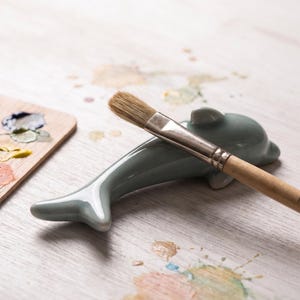 May include: A light grey ceramic dolphin-shaped brush rest with a smooth, glossy finish. A wooden paintbrush with a silver metal ferrule and light brown bristles is resting horizontally across the back of the dolphin figure.