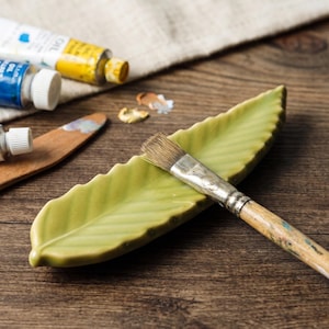 May include: A light green ceramic leaf-shaped brush rest with a textured surface and a central vein. A wooden-handled paintbrush with metal ferrule and light brown bristles rests across the center of the leaf, angled from the top right to bottom left.