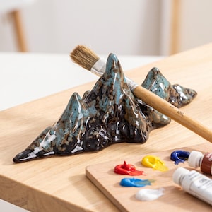 Könnte beinhalten: Eine Pinselablage aus Keramik in Form eines Gebirgszuges mit einer glänzenden Glasur in Dunkelbraun, Schwarz und Türkis. Das Stück weist mehrere Gipfel und Täler auf, die dazu dienen, einen Holzpinsel mit hellem Griff abzulegen.