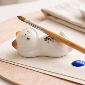 Könnte beinhalten: Eine weiße, katzenförmige Pinselablage aus Keramik mit braunen Ohrspitzen und einem lächelnden Gesicht. Die Katze trägt schwarze japanische Kalligrafie auf dem Rücken und stützt einen Pinsel mit Holzgriff und braunen Borsten ab.