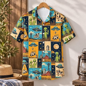 Könnte beinhalten: Ein kurzärmeliges Hemd mit Knopfleiste und einem Patchwork-Muster aus grünen Außerirdischen, fliegenden Untertassen, Kühen, roten Scheunen und Windmühlen auf dunkelblauem und gelbem Grund. Das Hemd hat einen klassischen Kragen und weiße Knöpfe.
