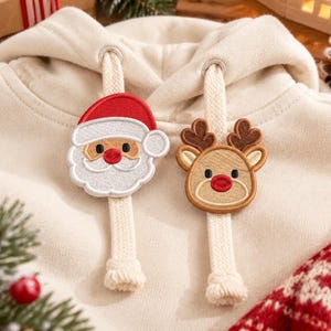May include: A cream-coloured hooded sweatshirt featuring two thick, flat drawstrings. Attached to each drawstring is a circular embroidered patch: one depicts a smiling Santa Claus with a red hat, and the other shows a brown reindeer with a red nose.