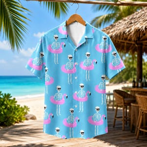 May include: A light blue short-sleeve button-down shirt featuring an all-over print of white skeletons wearing black sunglasses and floating in bright pink flamingo-shaped inner tubes. The shirt has a classic collar and a front button closure.
