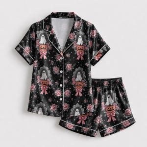 May include: A black pajama set featuring a short-sleeve button-down shirt and matching shorts. The fabric displays a repeating pattern of white ghosts sitting on orange pumpkins, pink roses, and small white stars with white piping trim.