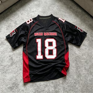 May include: A black short-sleeved sports jersey with red side panels and a V-neck collar. The front features the text "MEAN MACHINE" in red letters above the large white number "18" with a red outline. The number "18" is also on the shoulders.