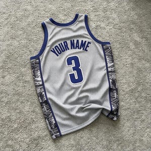 May include: A grey sleeveless basketball jersey featuring a dark blue trim around the neck and armholes. The back displays the text YOUR NAME in dark blue letters above the number 3 in dark blue with a white outline. Black and white side panels.