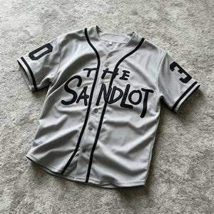 Op de afbeelding: Een lichtgrijs honkbalshirt met zwarte biezen langs de knoopsluiting en mouwen. De voorzijde is voorzien van de zwarte tekst "THE SANDLOT" in een schrijflettertype. Het getal "30" is in het zwart op beide mouwen gedrukt in een bloklettertype.