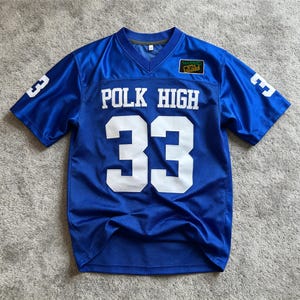 Puede incluir: Una camiseta azul con cuello en V, texto blanco POLK HIGH y el número 33 en la parte delantera. El número 33 también está impreso en ambas mangas. Un pequeño parche rectangular en el pecho presenta el texto MARRIED WITH CHILDREN en amarillo.