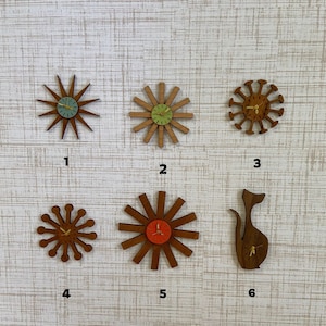 May include: A collection of six wooden wall clocks in various mid-century modern designs. The clocks feature starburst, sunburst, and atomic shapes with colored clock faces. The clock faces are in shades of green, orange, and brown.