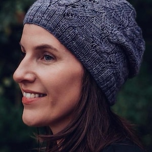 Könnte beinhalten: Eine Frau trägt eine dunkelgraue Strickmütze mit einem Zopfmuster. Die Mütze ist locker und hat eine lockere Passform. Die Frau hat lange dunkle Haare und lächelt.