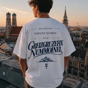Könnte beinhalten: Ein weißes T-Shirt mit kurzen Ärmeln und dunkelblauem Text auf der Rückseite, der WE DONT SAY VERFLIXT NOCHMAL WE SAY GREIZGRUZEFIX NUMMOINEI lautet, ergänzt durch eine grafische Darstellung eines Berggipfels und ein kleines rautenförmiges Symbol.