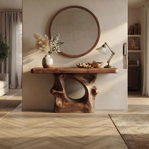 May include: A wooden console table with a sculptural, organic base featuring a central cutout. The table has a smooth, rectangular top and is positioned against a wall beneath a large, round mirror with a thin brown frame in a bright room.