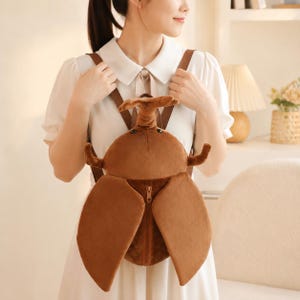 May include: A brown plush beetle backpack featuring a large horn, two smaller side horns, and two large wing-shaped panels that cover a central vertical zipper. The backpack includes two brown shoulder straps attached to the top and bottom.