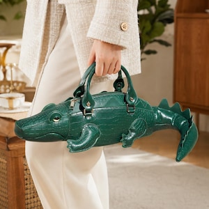 Può includere: Una borsa a mano verde scuro a forma di coccodrillo con una superficie testurizzata e goffrata che imita la pelle di rettile. La borsa presenta una chiusura a zip sulla parte superiore, due manici robusti e una coda prominente.