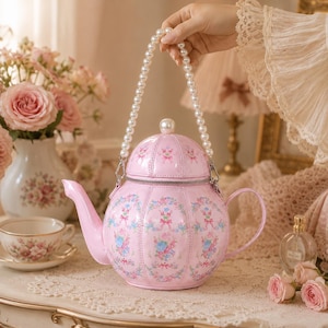 Peut inclure: Un sac à main rose clair en forme de théière orné d'un motif floral avec des fleurs bleues et roses, une fermeture éclair argentée et une anse en perles blanches. Le sac possède un bec verseur courbé et une anse latérale, imitant une théière.