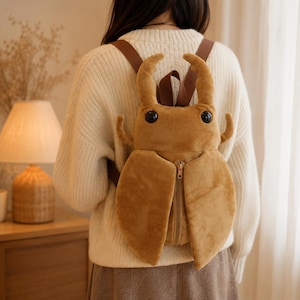 May include: A tan plush backpack shaped like a stag beetle with large mandibles, two black circular eyes, and a central vertical zipper closure. The bag features brown adjustable shoulder straps and is crafted from a soft, velvety fabric material.