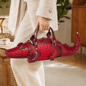 May include: A deep red handbag shaped like a crocodile with a textured, embossed surface. The bag features a zip closure, two top handles, a pointed tail, and a single visible yellow and black eye detail on the side of the crocodile head.
