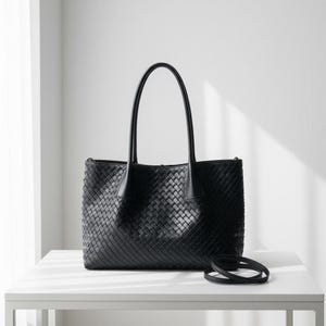 May include: A black tote bag featuring an intricate woven pattern across the entire exterior. The bag includes two tall, slender shoulder straps attached to the top edge and a separate, thin, coiled black strap resting on the surface below.