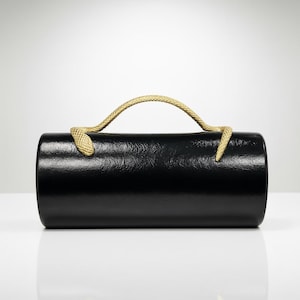 Può includere: Una borsa a mano cilindrica nera con una finitura lucida e testurizzata. La borsa presenta un manico singolo a forma di serpente color oro, con la testa e la coda del serpente fissate sulla parte superiore del cilindro, creando un arco.