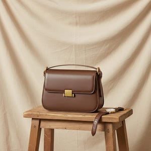Könnte beinhalten: Eine glatte, dunkelbraune Ledertasche mit strukturierter rechteckiger Form, einem Tragegriff und einem goldfarbenen Metallverschluss. Eine braune Lederarmbanduhr mit weißem Zifferblatt und goldfarbenen Akzenten liegt auf der Holzoberfläche.
