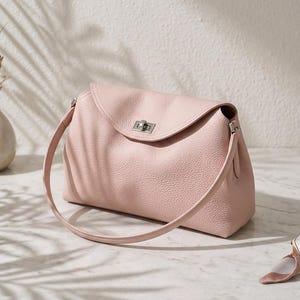 Könnte beinhalten: Eine hellrosa Handtasche mit strukturierter Lederoberfläche, ausgestattet mit einem geschwungenen Klappenverschluss, der durch einen silberfarbenen Metalldrehverschluss gesichert ist, sowie einem passenden dünnen Schulterriemen.
