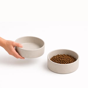 Hand lifting a stoneware ceramic pet bowl in oatmeal beside a matching bowl of dry food, showing the size of the MOTO dog and cat bowl in the hand