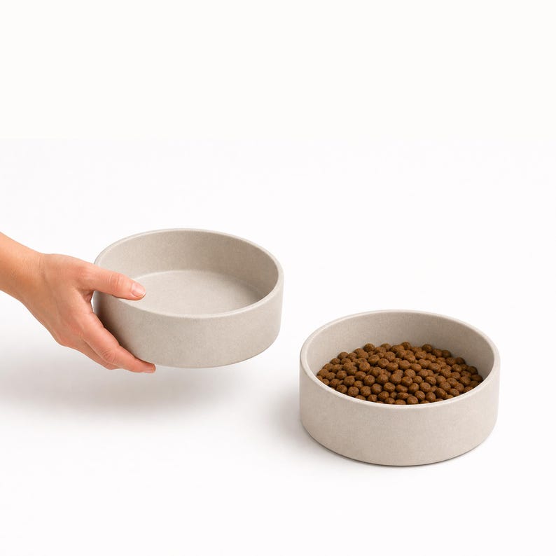 Hand lifting a stoneware ceramic pet bowl in oatmeal beside a matching bowl of dry food, showing the size of the MOTO dog and cat bowl in the hand