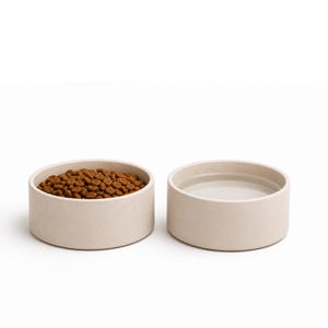 Two matching ceramic dog and cat bowls in oatmeal speckled stoneware side by side, one with dry food and one with water, on a white background