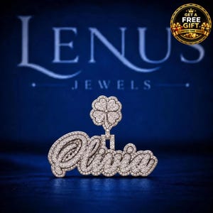 May include: A silver-toned pendant featuring the name Olivia in cursive script, encrusted with numerous small clear stones. A four-leaf clover charm, also covered in clear stones, is attached to the top of the nameplate.