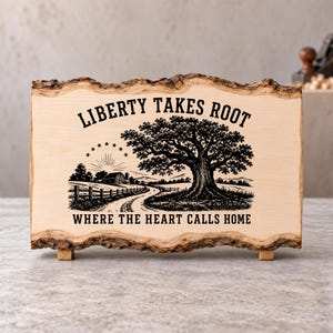 May include: A rectangular light-colored wooden plaque with natural bark edges, featuring a black ink illustration of a large oak tree, a barn, a fence, and a sunburst with stars. The text reads: LIBERTY TAKES ROOT WHERE THE HEART CALLS HOME.