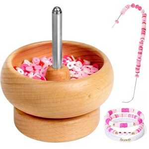 May include: A wooden bead spinner bowl with a central metal spindle containing pink, white, and red clay disc beads. A curved metal needle holds a string of matching beads, and three finished bracelets with pink and white beads are shown.