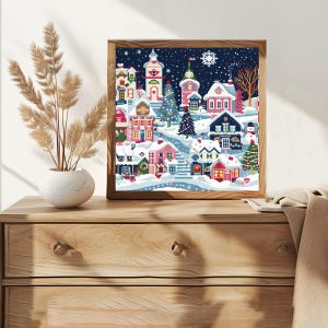 May include: A square framed wall art piece featuring a colourful winter village scene with snow-covered houses, decorated Christmas trees, and a snowman under a dark blue night sky filled with white snowflakes and a large white snowflake icon.