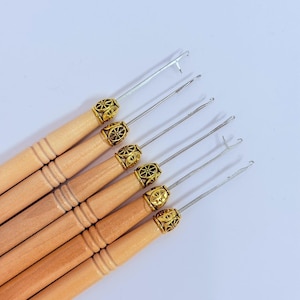 May include: A set of six latch hook tools featuring light-colored wooden handles with three horizontal grooves. Each tool has a metallic shaft ending in a latch hook mechanism, secured by a decorative gold-toned metal ferrule with a wheel pattern.
