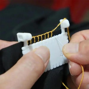 May include: A white plastic sewing tool with notched edges used for spacing stitches, featuring yellow thread looped through the notches. A silver sewing needle is positioned near the tool, which is placed against a piece of black fabric.