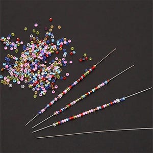 May include: Four long, thin, silver-colored metal needles. Three of the needles are threaded with a variety of small, multi-colored glass beads in shades of red, blue, green, yellow, and pink. A loose pile of these same small beads sits nearby.