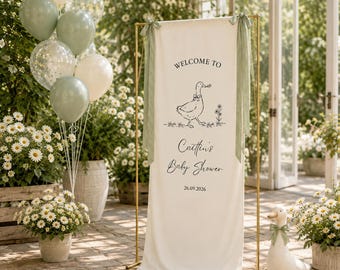 May include: A vertical cream-colored fabric banner on a gold metal stand, featuring a black line drawing of a goose, the text "WELCOME TO", "Caitlin's Baby Shower", and "26.09.2026". The banner is tied with sage green ribbons at the top corners.