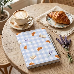 May include: A hardback notebook featuring a light blue and white gingham pattern decorated with small illustrations of golden brown croissants, open books, and sprigs of purple lavender, accompanied by a silver metal pen resting on the cover.