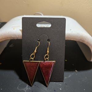 May include: A pair of dangle earrings featuring dark red, shimmering triangular resin pendants set in gold-toned metal frames. The earrings hang from gold-toned fishhook wires and are displayed on a rectangular black cardboard jewelry card.