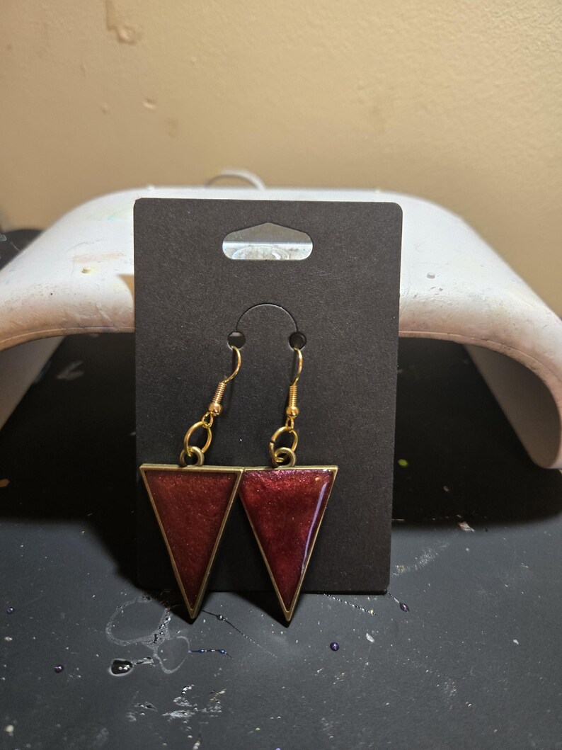 May include: A pair of dangle earrings featuring dark red, shimmering triangular resin pendants set in gold-toned metal frames. The earrings hang from gold-toned fishhook wires and are displayed on a rectangular black cardboard jewelry card.