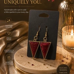 May include: A pair of gold-toned triangle earrings featuring a deep red, glittery resin inlay. The earrings are suspended from gold-toned metal hooks and are displayed on a black rectangular card. The design is geometric and minimalist in shape.