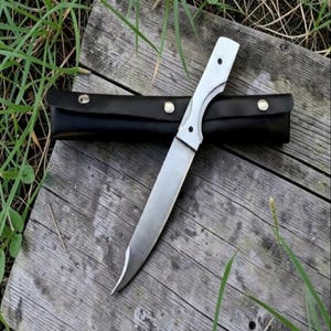 Puede incluir: Un cuchillo de hoja fija con una hoja de metal plateado y un mango a juego con dos remaches visibles. El cuchillo está colocado en diagonal sobre una funda de cuero negro que tiene tres broches de presión de metal plateado a lo largo.