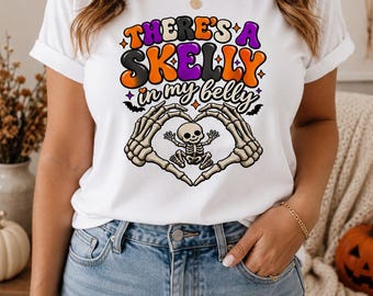 May include: A white short-sleeved t-shirt featuring a graphic print with the text "THERE'S A SKELLY in my belly" in orange, black, and purple. Below the text, two skeletal hands form a heart shape around a small cartoon skeleton figure.