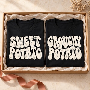 Puede incluir: Dos camisetas negras dobladas dentro de una caja de cartón. La camiseta de la izquierda presenta el texto color crema "SWEET POTATO" en una fuente ondulada y desgastada. La camiseta de la derecha presenta el texto "GROUCHY POTATO" igual.