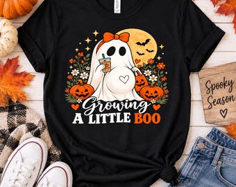 May include: A black t-shirt featuring a graphic of a ghost wearing an orange bow, holding a drink and a pumpkin bucket. The design includes pumpkins, flowers, bats, a moon, and the text "Growing A LITTLE BOO" in white and orange lettering.