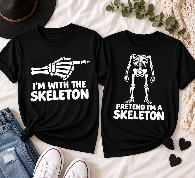 May include: Two black short-sleeved t-shirts. The left shirt features a white skeleton hand pointing right with the text I'M WITH THE SKELETON. The right shirt displays a white skeleton torso and leg graphic with the text PRETEND I'M A SKELETON.