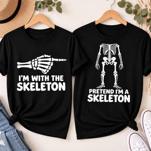 May include: Two black short-sleeve t-shirts. The left shirt features a white skeleton hand pointing right with the text I'M WITH THE SKELETON. The right shirt displays a white skeleton torso and legs with the text PRETEND I'M A SKELETON.