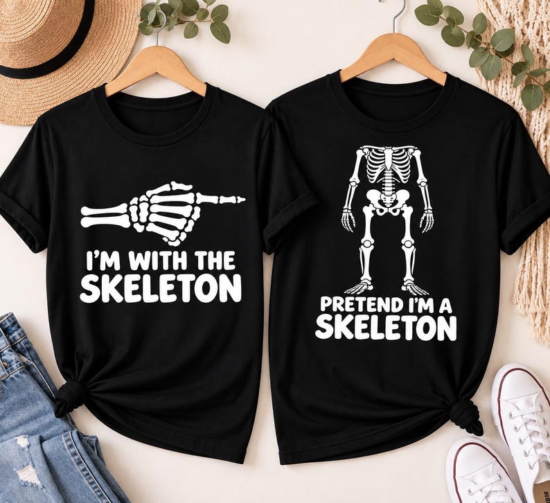 May include: Two black short-sleeve t-shirts. The left shirt features a white skeleton hand pointing right with the text I'M WITH THE SKELETON. The right shirt displays a white skeleton torso and legs with the text PRETEND I'M A SKELETON.