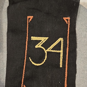Peut inclure: Un écusson rectangulaire en tissu noir présentant le nombre 34 brodé avec un fil jaune doré. Le nombre est centré et entouré d'une fine bordure rectangulaire cousue avec un fil orange brûlé, agrémentée d'accents décoratifs aux coins.