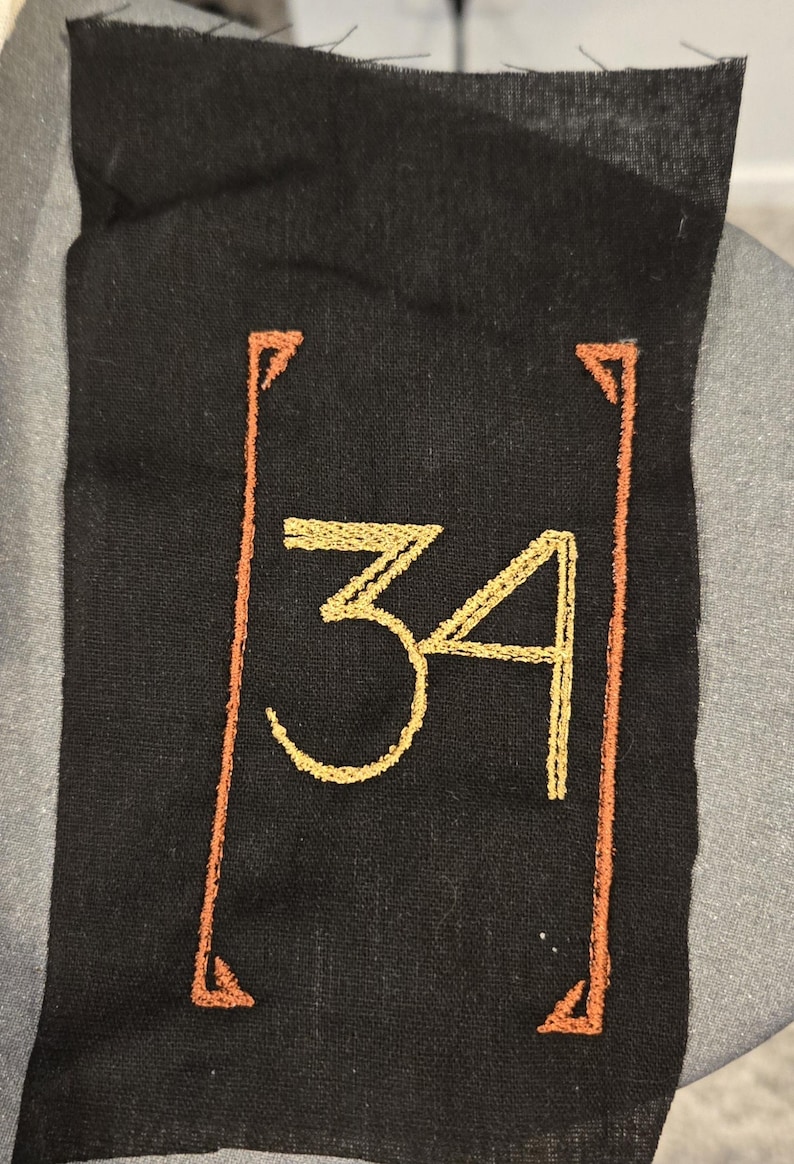 May include: A rectangular black fabric patch featuring the number 34 embroidered in a golden yellow thread. The number is centred and enclosed within a thin, rectangular border stitched in a burnt orange thread with decorative corner accents.